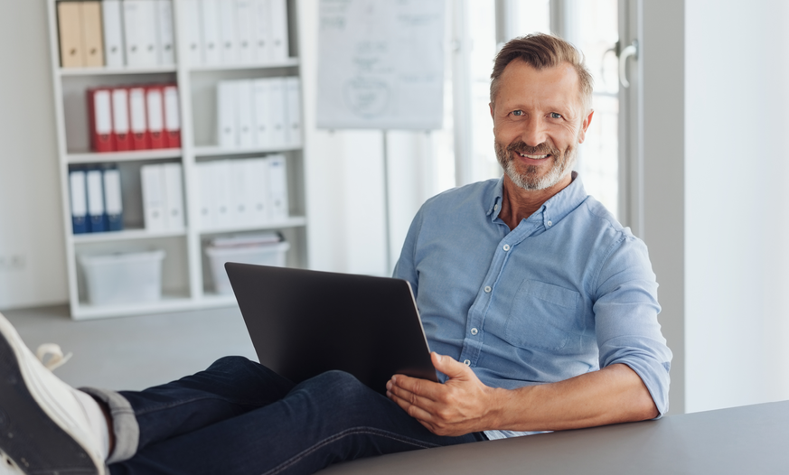 Mann mit hellblauem Hemd sitzt entspannt in Büro an einem Schreibtisch und hat Laptop auf dem Schoß Mann mit Laptop auf dem Schoß